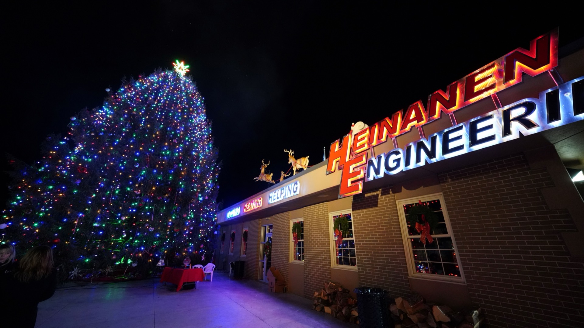 The Heinanen Engineering building lit up at night in South Lyon, Michigan, with its orange neon sign and the community holiday tree